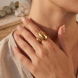 Peut inclure: Une bague dorée avec une grande forme rectangulaire arrondie et un petit accent circulaire. La bague est portée au doigt, reflétant la lumière. Ce bijou est une bague de caractère.