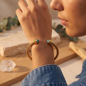 Könnte beinhalten: Ein goldfarbenes Armband in Manschettenform mit gedrehtem Metallband. Das offene Armband ist an den Enden mit zwei abgerundeten, polierten goldfarbenen Kappen versehen, die jeweils in der Mitte einen kleinen, glatten, hellblauen Türkisstein tragen.