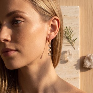 Peut inclure: Une boucle d'oreille créole dorée avec un design pendant vertical composé de trois petites pierres transparentes de taille rectangulaire alignées. La boucle d'oreille est portée au lobe, présentant une finition métallique polie et des pierres facettées.