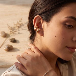 Peut inclure: Un ear cuff doré au design torsadé et multicouche qui s'enroule autour du cartilage de l'oreille. La pièce présente une finition métallique polie et se compose de deux bandes entrelacées créant un aspect arrondi et continu.