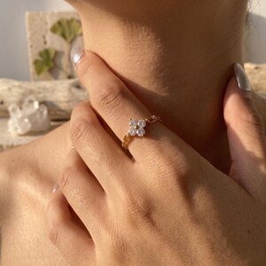 Peut inclure: Une bague dorée ornée d'un motif floral à quatre pétales orné de pierres claires. La bague est montée sur un anneau en forme de chaîne. La bague est portée au doigt.