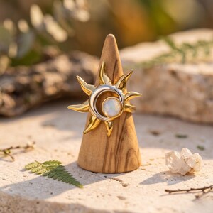 Peut inclure: Une bague dorée présentant un motif de rayons de soleil pointus entourant un croissant de lune argenté central et une pierre ronde blanche irisée. La bague est présentée sur un support conique en bois lisse.