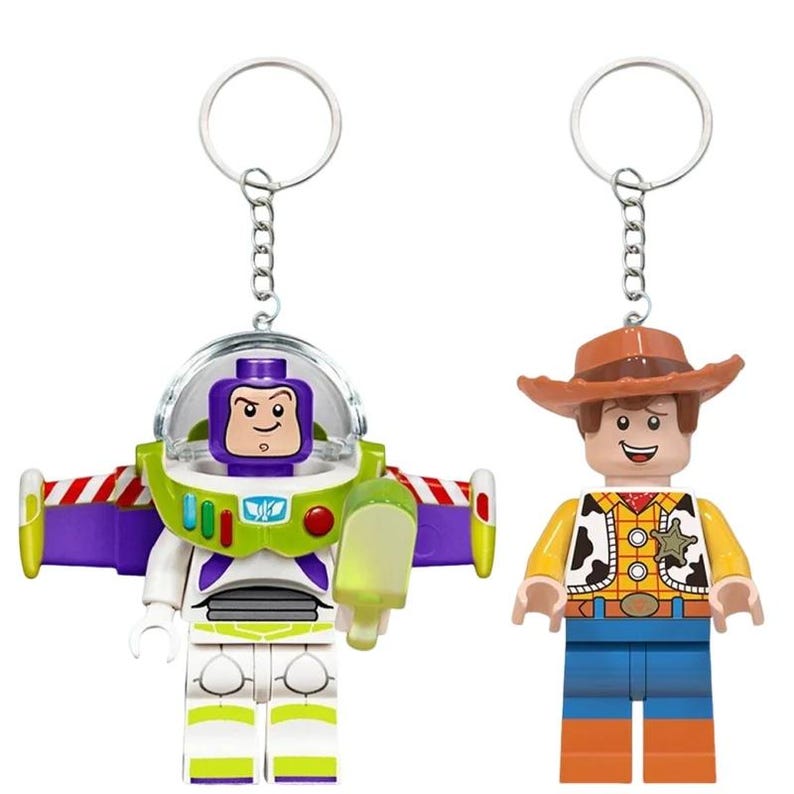 Puede incluir: Dos llaveros de personajes que incluyen una figura de guardi&aacute;n espacial con casco de c&uacute;pula transparente, alas moradas y detalles verdes, junto a una figura de vaquero con sombrero marr&oacute;n, camisa amarilla con chaleco de manchas y pantalones azules.