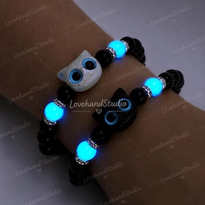 Puede incluir: Dos pulseras de cuentas con cuentas negras y detalles azules brillantes. Cada pulsera presenta un dije en forma de gato, uno blanco y otro negro, y una cuenta azul brillante con detalles plateados. Las pulseras están en una muñeca.