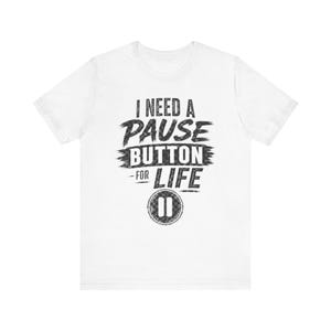 May include: White t-shirt featuring the phrase "I NEED A PAUSE BUTTON FOR LIFE" in a distressed black font. The design includes a pause button graphic. A casual wear item.