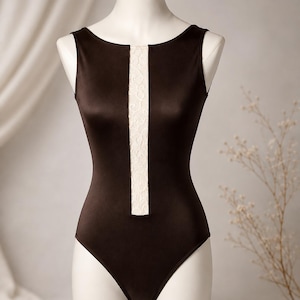 May include: A sleeveless dark brown bodysuit featuring a vertical cream-coloured lace panel running down the centre of the chest. The garment has a high, rounded neckline and a fitted silhouette, displayed on a mannequin torso.