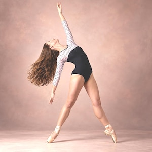 May include: A long-sleeved leotard featuring a solid black bodice and white sleeves adorned with a black polka dot pattern. The garment is worn by a dancer in a pose with one arm extended upward, paired with light pink satin ballet pointe shoes.