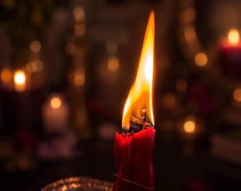 Könnte beinhalten: Eine hohe, rote Wachskerze mit einer hellen, flackernden Flamme an der Spitze. Die Kerze ist mit einem dünnen, weißen Faden umwickelt und steht in einem rechteckigen Behälter aus Aluminiumfolie, der das warme Licht des Feuers reflektiert.