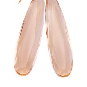 Puede incluir: Un par de pendientes colgantes dorados con cristales facetados en forma de lágrima de color melocotón. Los pendientes tienen un diseño sencillo y elegante, con un gancho de alambre dorado y un detalle enrollado en la parte superior de cada cristal.