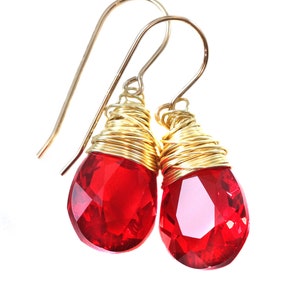 May include: A pair of red crystal teardrop earrings with gold wire wrapped around the crystal. The earrings have gold hooks.
