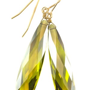 Può includere: Un paio di orecchini pendenti color oro con cristalli sfaccettati a goccia verde oliva. Gli orecchini hanno un semplice design a gancio, su sfondo bianco.
