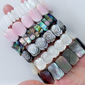 Op de afbeelding: Een stapel van zes armbanden gemaakt van verschillende soorten schelpen. De armbanden hebben verschillende ontwerpen, waaronder ovale, rechthoekige en roosvormige kralen. De kleuren omvatten wit, roze, zwart en iriserend groen.