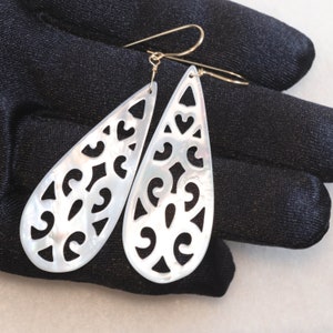 May include: A pair of white mother of pearl earrings with a delicate, intricate cut-out design. The earrings are teardrop shaped and have gold hooks.