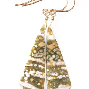 May include: A pair of gold-colored wire earrings with triangular, patterned stone pendants. The pendants feature a mix of green, white, and tan colors. The earrings are displayed against a white background.