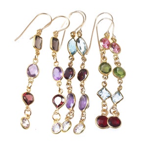 May include: Five gold dangle earrings featuring multi-colored faceted gemstones in various shapes including round, oval, pear, and square. Each earring is suspended from a gold hook and displays a unique vertical arrangement of colorful stones.
