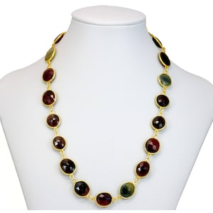 以下が含まれることがあります： 赤、緑、茶色の色合いの楕円形の宝石がついた金のチェーンネックレス。ネックレスは白いマネキンに飾られています。