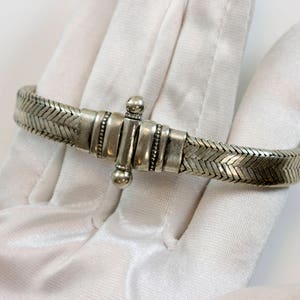 Puede incluir: Un brazalete trenzado de plata con un cierre de plata. El brazalete está sobre una superficie blanca.