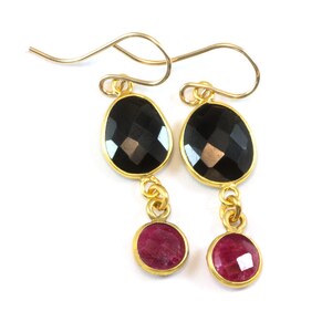 May include: A pair of gold-colored earrings with a black, faceted oval stone at the top and a smaller, round, red stone at the bottom. The stones are connected by a gold chain. The earrings have a hook closure.