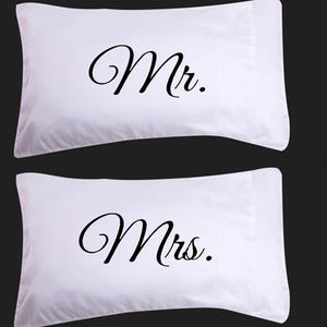Puede incluir: Dos fundas de almohada blancas con texto negro en cursiva. Una funda de almohada dice "Mr." y la otra "Mrs.". Las fundas de almohada son rectangulares y tienen un diseño simple y elegante.