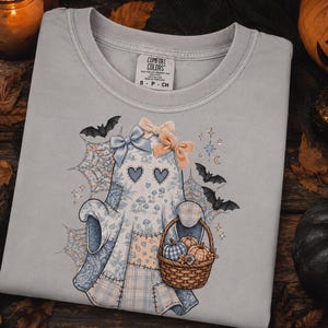 May include: A light grey t-shirt featuring a graphic of a ghost with heart-shaped eyes, wearing blue and peach bows. The ghost is decorated with a blue and white mushroom pattern and holds a woven basket filled with small pumpkins and gourds.