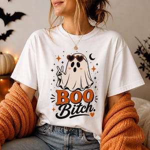 Op de afbeelding: Wit T-shirt met een Halloween-thema. Het ontwerp toont een cartoongeest met een hartvormige zonnebril en een vredesteken, met de woorden "BOO Bitch" in oranje en zwart. Het shirt maakt deel uit van een Halloween-collectie.
