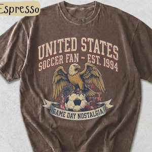 May include: Espresso-colored t-shirt featuring a graphic of an eagle perched on a soccer ball, with the text "UNITED STATES SOCCER FAN - EST. 1994" and "GAME DAY NOSTALGIA". The shirt has a vintage, distressed look.