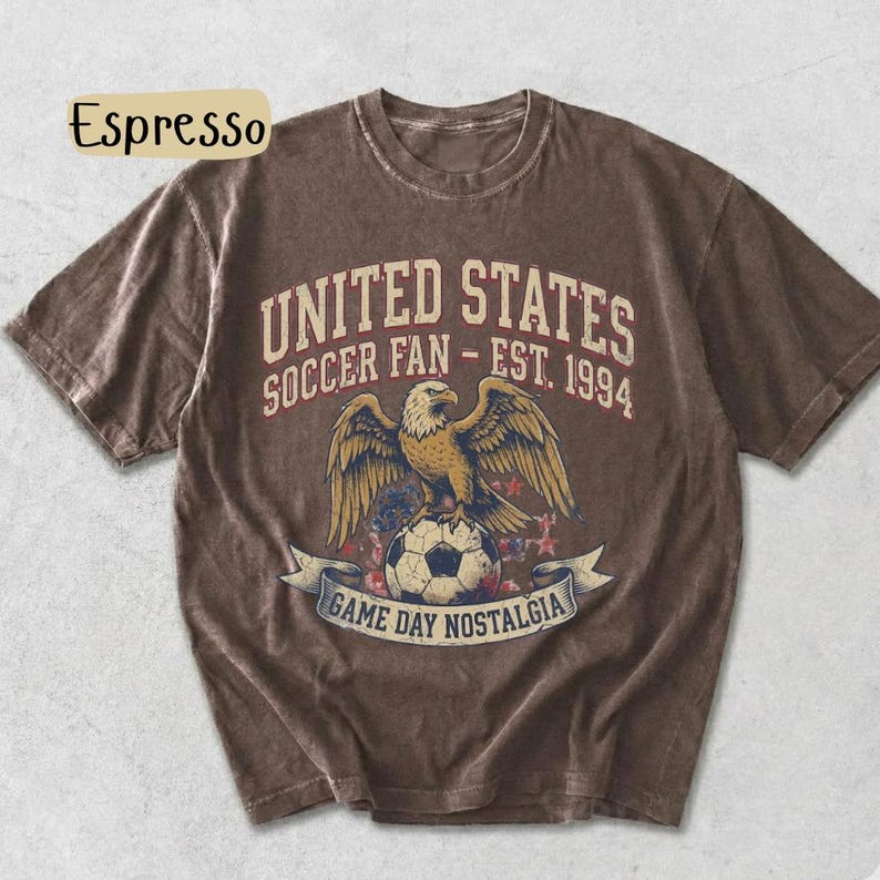 May include: Espresso-colored t-shirt featuring a graphic of an eagle perched on a soccer ball, with the text "UNITED STATES SOCCER FAN - EST. 1994" and "GAME DAY NOSTALGIA". The shirt has a vintage, distressed look.