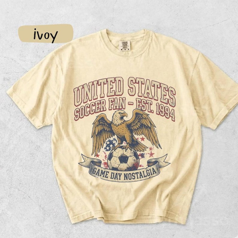 May include: Ivory-colored t-shirt with a graphic featuring an eagle perched on a soccer ball. The text reads "UNITED STATES SOCCER FAN - EST. 1994" above and "GAME DAY NOSTALGIA" below. The shirt has a crew neck and short sleeves.