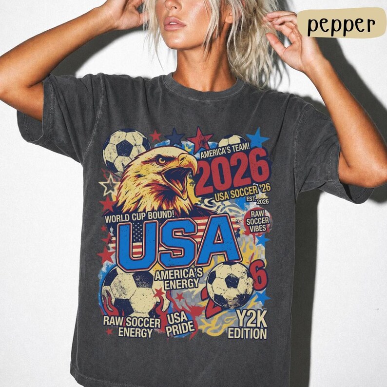 K&ouml;nnte beinhalten: Dunkelgraues T-Shirt mit einem Grafikdesign mit einem Wei&szlig;kopfseeadler, Fu&szlig;b&auml;llen und dem Text "USA 2026". Das Design enth&auml;lt die S&auml;tze "World Cup Bound!" und "America's Team!" im Vintage-Stil. Das T-Shirt hat einen Rundhalsausschnitt.