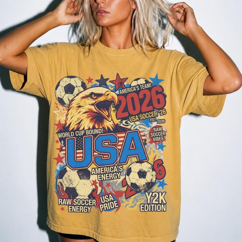 K&ouml;nnte beinhalten: Senfgelbes T-Shirt mit einem Grafikdesign, das einen Adler, Fu&szlig;b&auml;lle und den Text "USA 2026" zeigt. Das Design enth&auml;lt Sterne und die S&auml;tze "World Cup Bound", "America's Team" und "Raw Soccer Vibes".