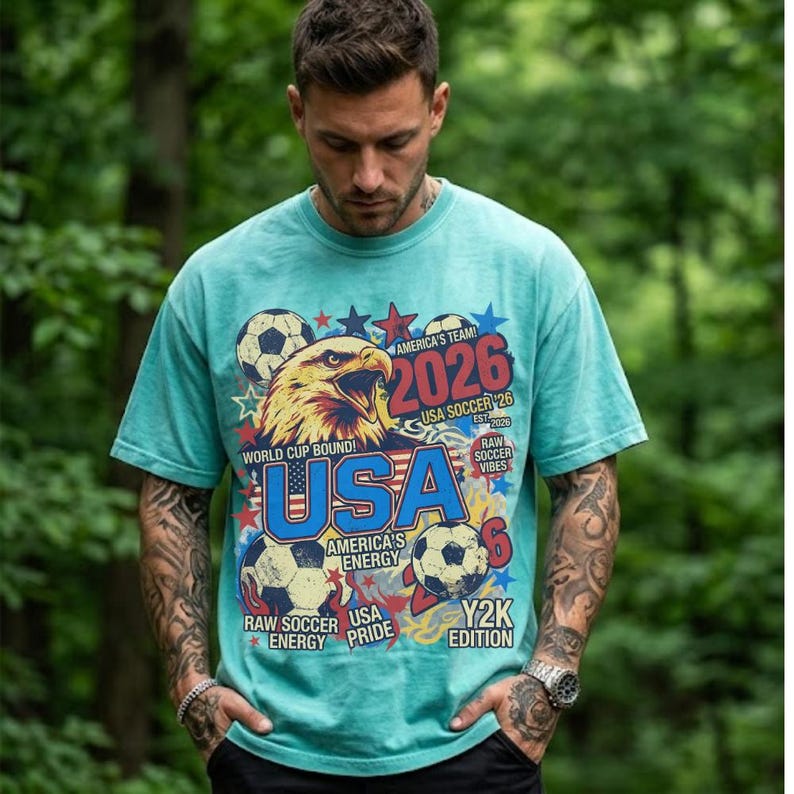 K&ouml;nnte beinhalten: T&uuml;rkisfarbenes T-Shirt mit einem Grafikdesign mit einem Adler, Fu&szlig;b&auml;llen und dem Text "USA 2026". Das Design enth&auml;lt die S&auml;tze "World Cup Bound!" und "America's Energy". Das Shirt hat einen Vintage-Y2K-Stil.
