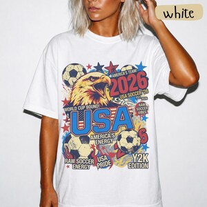 K&ouml;nnte beinhalten: Wei&szlig;es T-Shirt mit einem farbenfrohen Grafikdesign, das einen Adler, Fu&szlig;b&auml;lle und den Text "USA 2026" zeigt. Das Design enth&auml;lt auch die Texte "America's Team!" und "World Cup Bound!". Ein l&auml;ssiges, sportliches T-Shirt.