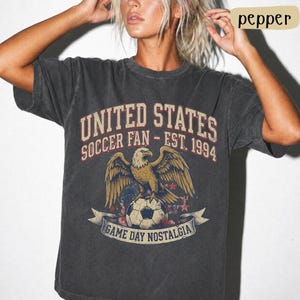 May include: Dark gray t-shirt with a vintage graphic. The design features an eagle perched on a soccer ball, with text that reads "UNITED STATES SOCCER FAN - EST. 1994" and "GAME DAY NOSTALGIA".