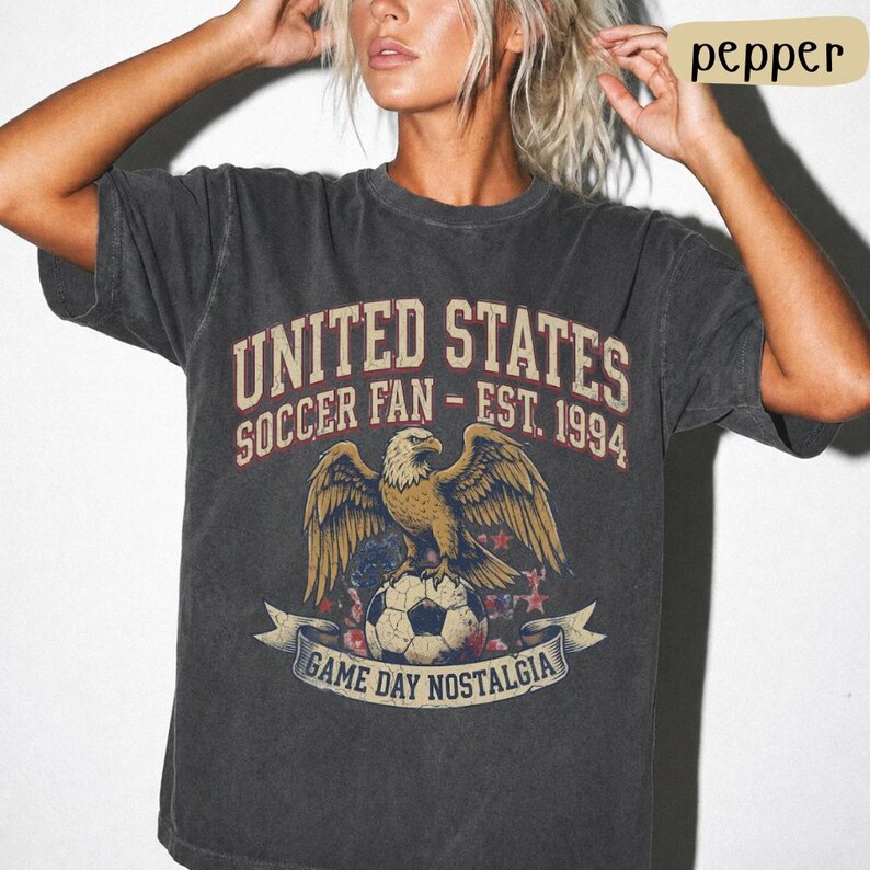May include: Dark gray t-shirt with a vintage graphic. The design features an eagle perched on a soccer ball, with text that reads "UNITED STATES SOCCER FAN - EST. 1994" and "GAME DAY NOSTALGIA".
