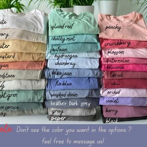 K&ouml;nnte beinhalten: Gestapelte T-Shirts in verschiedenen Farben, darunter Wei&szlig;, Elfenbein und Senf. Weitere Farben sind Island Reef, Kreideminze, Pfirsich und Crunchberry. Das Bild enth&auml;lt auch den Text "Hinweis: Sehen Sie die gew&uuml;nschte Farbe nicht in den Optionen? Kontaktieren Sie uns gerne!"