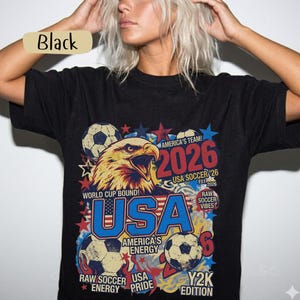K&ouml;nnte beinhalten: Schwarzes T-Shirt mit einem Grafikdesign, das einen Adler, Fu&szlig;b&auml;lle und den Text "USA 2026" zeigt. Das Design enth&auml;lt den Text "World Cup Bound!" und "America's Team!" in Rot, Wei&szlig; und Blau.