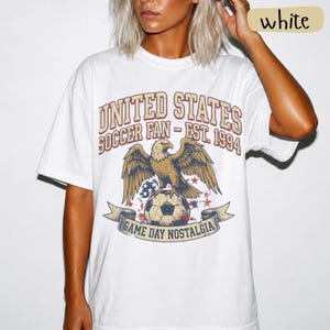 May include: White t-shirt with a vintage-style graphic featuring an eagle perched on a soccer ball. The text reads "UNITED STATES SOCCER FAN - EST. 1994" and "GAME DAY NOSTALGIA". The shirt has short sleeves and a relaxed fit.