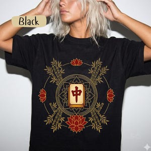May include: Black t-shirt with a gold and red graphic. The design features bamboo, lotus flowers, and a central tile with Chinese characters. The word "Black" is printed on a tan label.