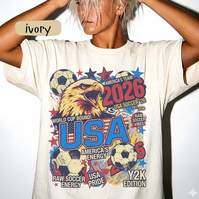 K&ouml;nnte beinhalten: Elfenbeinfarbenes T-Shirt mit einem Grafikdesign mit einem Adler, Fu&szlig;b&auml;llen und dem Text "USA 2026". Das Design enth&auml;lt Sterne und die S&auml;tze "World Cup Bound", "America's Team" und "Raw Soccer Vibes".