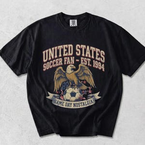 May include: Black t-shirt with a graphic featuring a golden eagle perched on a soccer ball. Text reads "UNITED STATES SOCCER FAN - EST. 1994" above and "GAME DAY NOSTALGIA" below. The design includes stars and a banner.