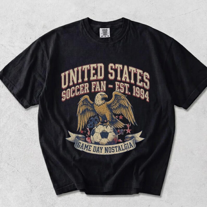 May include: Black t-shirt with a graphic featuring a golden eagle perched on a soccer ball. Text reads "UNITED STATES SOCCER FAN - EST. 1994" above and "GAME DAY NOSTALGIA" below. The design includes stars and a banner.