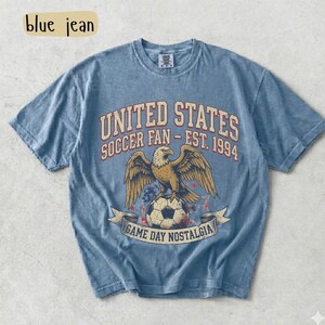 May include: Blue t-shirt with a graphic featuring an eagle perched on a soccer ball, with text reading "UNITED STATES SOCCER FAN - EST. 1994" and "GAME DAY NOSTALGIA". The shirt has a vintage, faded look.