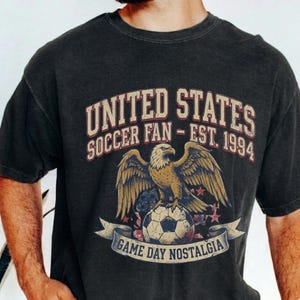 May include: Dark gray t-shirt with a distressed graphic. The graphic features a golden eagle perched on a soccer ball, with text that reads "UNITED STATES SOCCER FAN - EST. 1994" and "GAME DAY NOSTALGIA" on a banner. The shirt has short sleeves.