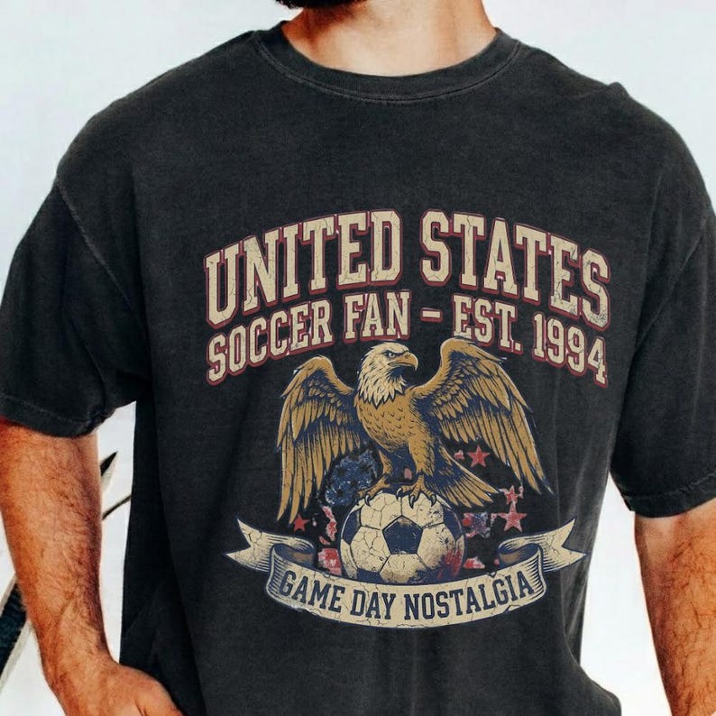 May include: Dark gray t-shirt with a distressed graphic. The graphic features a golden eagle perched on a soccer ball, with text that reads "UNITED STATES SOCCER FAN - EST. 1994" and "GAME DAY NOSTALGIA" on a banner. The shirt has short sleeves.
