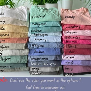 May include: A selection of stacked t-shirts in a variety of colours, such as white, ivory, and mustard. Other colours include island reef, chalky mint, peachy, and crunchberry. The image also includes text that says "note: Don't see the colour you want in the options? feel free to message us!"