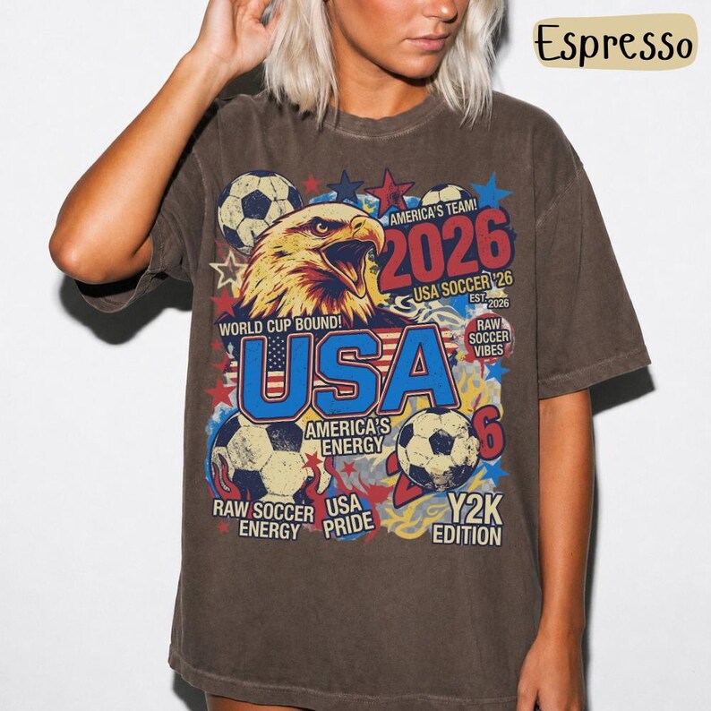 K&ouml;nnte beinhalten: Espressofarbenes T-Shirt mit einem Grafikdesign mit einem Adler, Fu&szlig;b&auml;llen und dem Text "USA 2026". Das Design enth&auml;lt die W&ouml;rter "World Cup Bound", "America's Team" und "Raw Soccer Vibes".