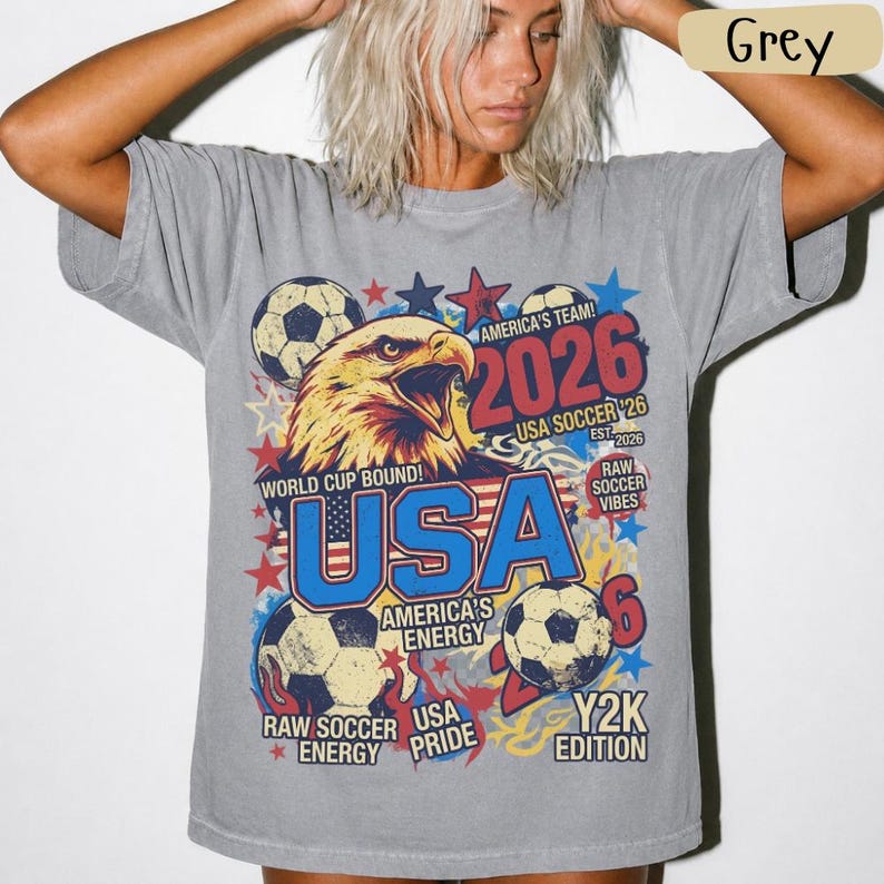 K&ouml;nnte beinhalten: Graues T-Shirt mit einem Grafikdesign mit einem Adler, Fu&szlig;b&auml;llen, Sternen und dem Text "USA 2026". Das Shirt enth&auml;lt die S&auml;tze "America's Team", "World Cup Bound" und "Raw Soccer Vibes".