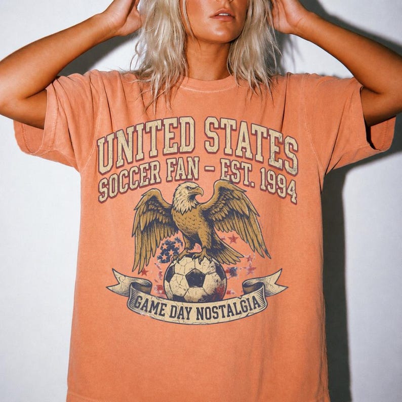 May include: Orange t-shirt with a distressed graphic. The graphic features a bald eagle perched on a soccer ball, with text that reads "UNITED STATES SOCCER FAN - EST. 1994" and "GAME DAY NOSTALGIA".