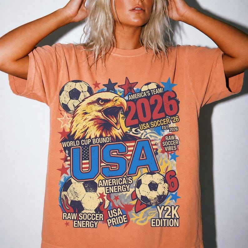K&ouml;nnte beinhalten: Orangefarbenes T-Shirt mit einem Grafikdesign, das einen Wei&szlig;kopfseeadler, Fu&szlig;b&auml;lle und den Text "USA 2026" zeigt. Das Design enth&auml;lt die S&auml;tze "World Cup Bound", "America's Energy" und "Y2K Edition".
