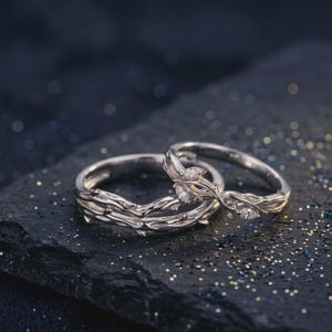 Puede incluir: Dos anillos de plata con un diseño inspirado en la naturaleza. Uno presenta una textura similar a una rama, mientras que el otro tiene una decoración de hojas y piedras preciosas. Los anillos están sobre un fondo oscuro y moteado.